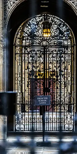 An intricately designed wrought iron gate leading into a school building, featuring a sign that reads 'FROM THE SCHOOL GATES'.